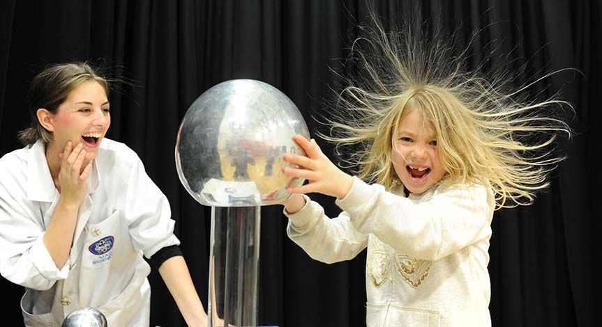 Girl touching a plasma ball causing her hair to to stand up while her mad science instructor looks on.
