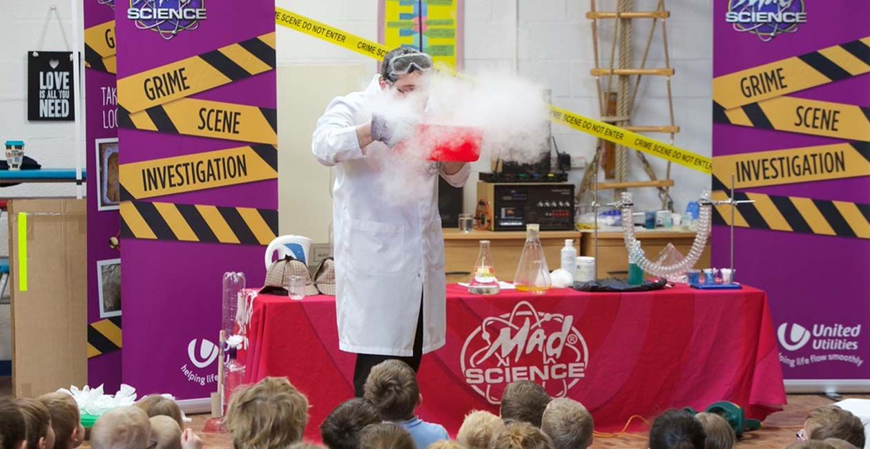 Mad scientist blowing into a red bowl with smoke coming out of it while kids watch